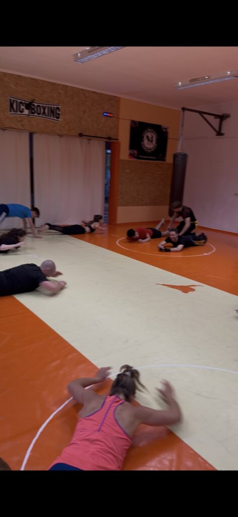 Teilnehmer eines Kickbox-Trainings machen Bodenübungen auf einer orangefarbenen Kampfsportmatte in einer Trainingshalle.